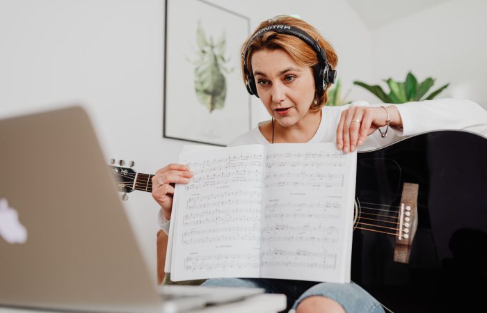 Woman teaching guitar online, presenting sheet music via video call with laptop setup.