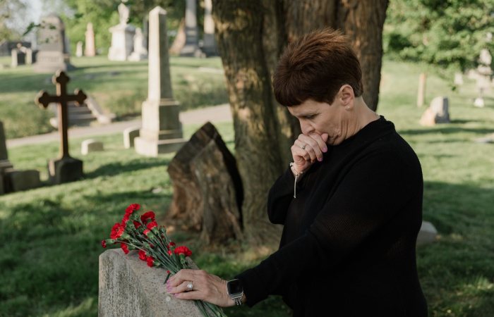 Senior woman in black at a cemetery, grieving by a gravestone with red flowers.